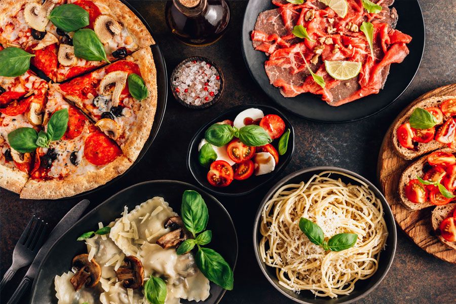 Full table of italian meals on plates Pizza, pasta, ravioli, carpaccio. caprese salad and tomato bruschetta on black background. Top view